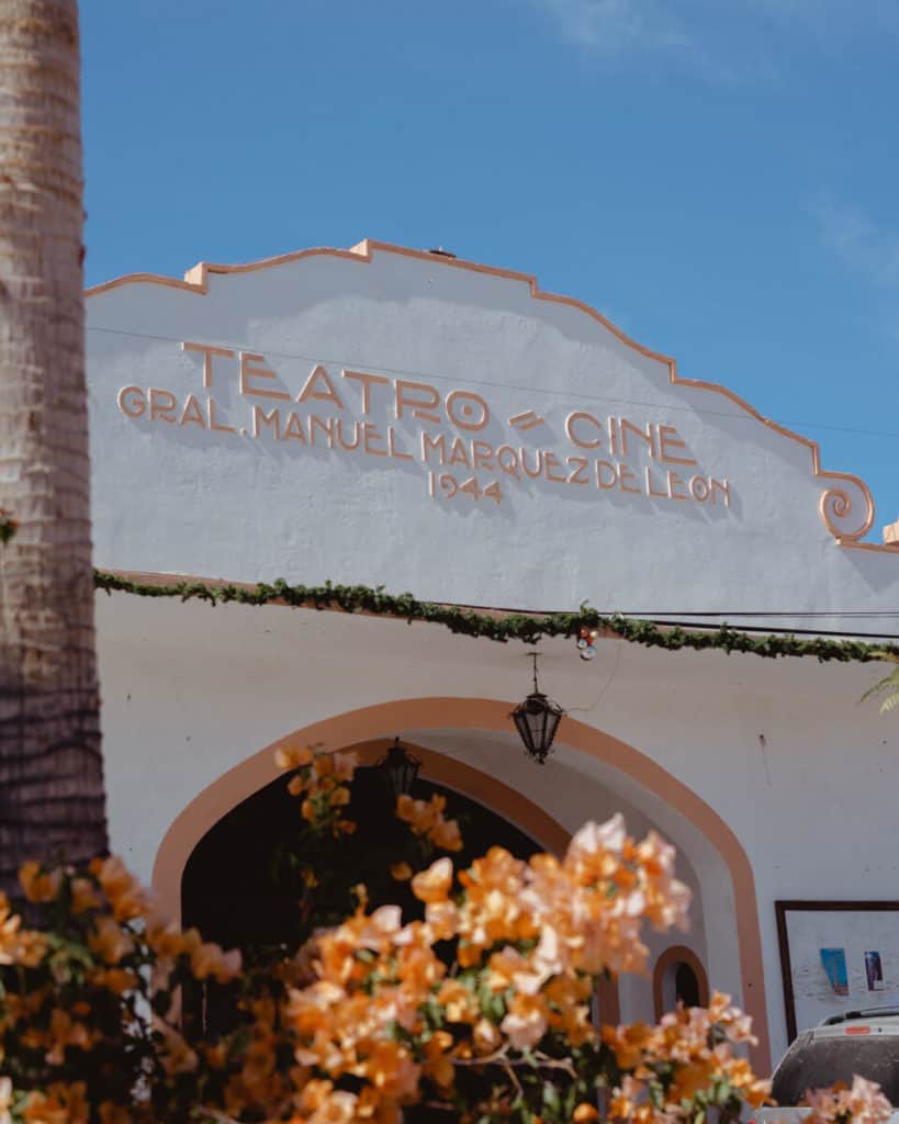 The white and peach facade of the historic Teatro-Cine General Manuel Márquez de León from 1944 is framed by flowering vines and palm trees in Todos Santos, Mexico.