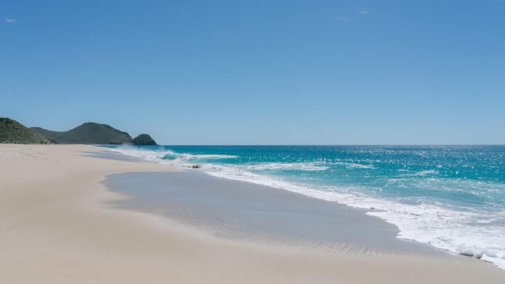 A pristine, empty stretch of golden sand meets turquoise waves under a clear blue sky at a peaceful beach near Todos Santos, Mexico.