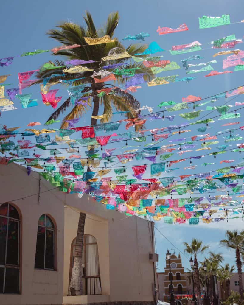 Colorful papel picado flags hang above a sunlit street lined with palm trees and colonial buildings, with the iconic Todos Santos church visible in the background in Todos Santos, Mexico.