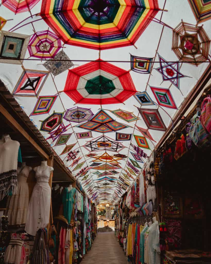 A colorful handmade yarn canopy hangs above a narrow artisan market in Todos Santos, Mexico, lined with traditional dresses, hats, and handcrafted goods.