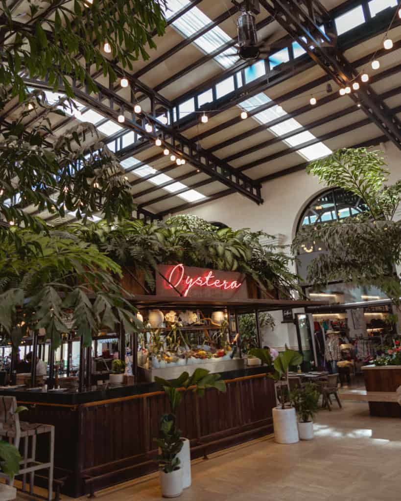 The lush, plant-filled interior of Oystera in Todos Santos features a stylish wooden bar with a neon sign and hanging globe lights under a vaulted ceiling.
