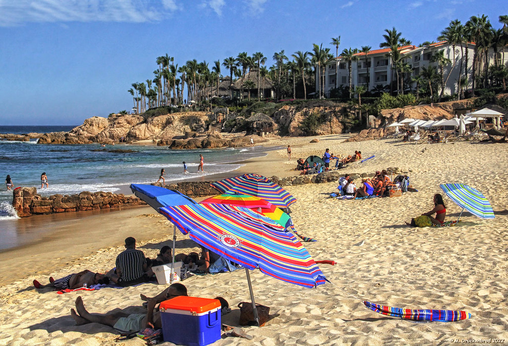 Beachgoers and colorful umbrellas at the shoreline identified by the photographer as Cabo Bello